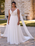 A woman in a flowing white wedding gown with lace details and shoulder drapes.